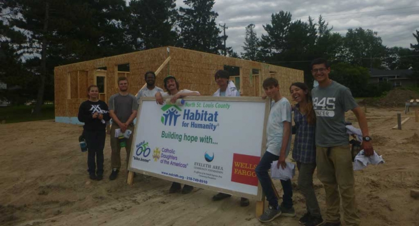 A group of people pose for a photo around a habitat for humanity sign. Behind them is the wooden frame of a house. 